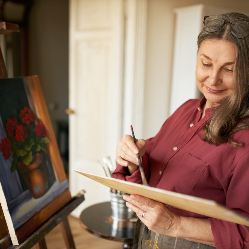 Image of creative skillful middle aged woman artist enjoying her hobby, standing next to easel at home, mixing oil pigments on palette using brush, working on painting of flower pot, smiling