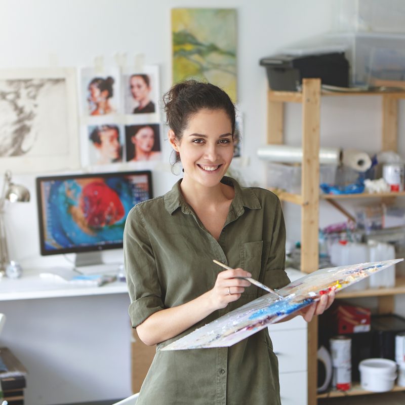 Indoor shot of beautiful brunette female painter wearing shirt, holding paint brush in hands standing near easel, creating masterpiece, smiling pleasantly into camera while being glad to paint
